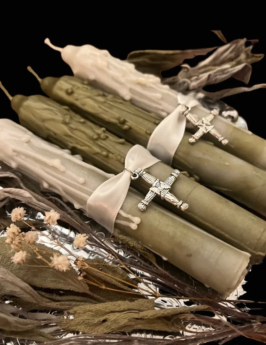 Cream and sage-green taper candles tied with ribbon and Brigid's cross charms among dried flowers