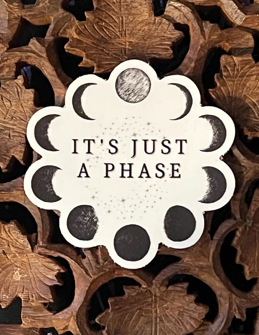 Sticker ringed with moon phases reading 'It's just a phase' on a carved wooden surface