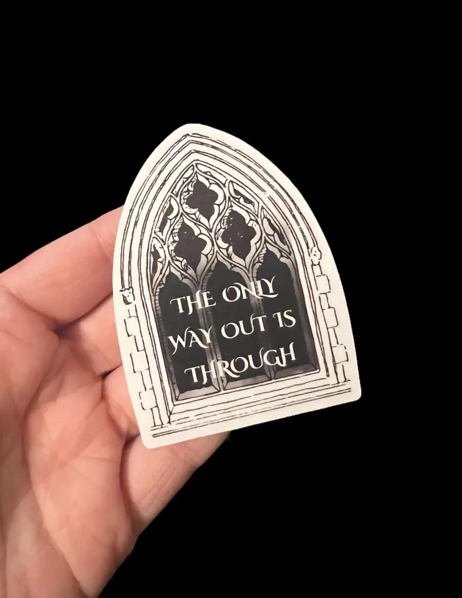 Hand holding an arched gothic-window sticker reading 'The only way out is through'